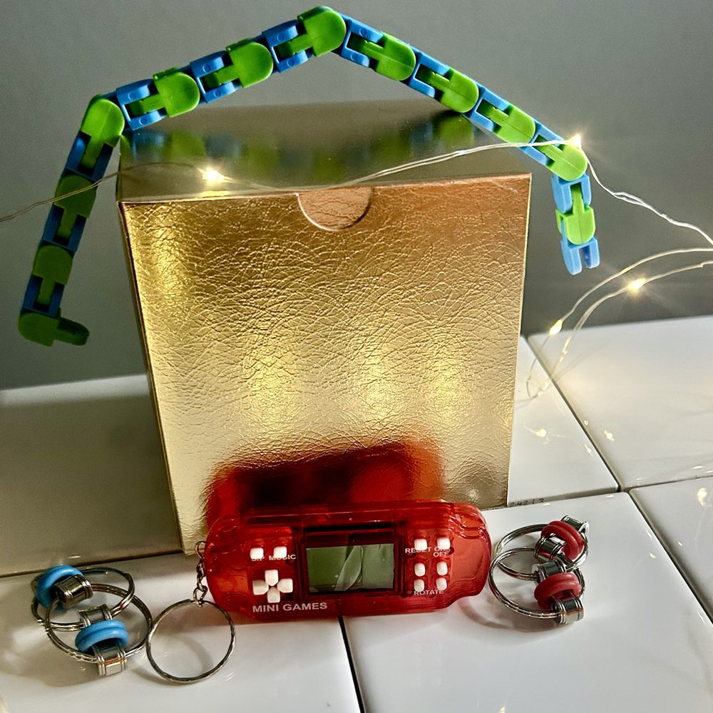 A gold gift box surrounded by fidget toys: a green and blue wacky track, a red mini handheld game, and two flippy chain key rings, with fairy lights